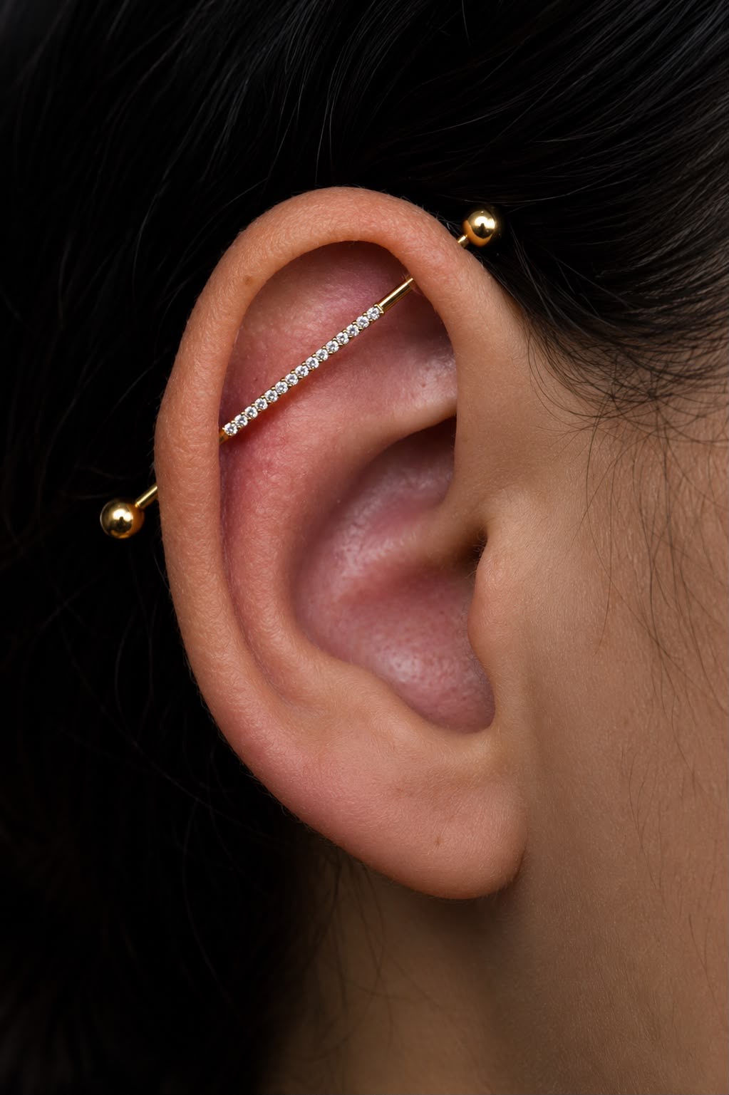 Piercing industrial com barra reta dourada cravejada de zircônias atravessando a cartilagem superior da orelha — Luquini Estética Auricular